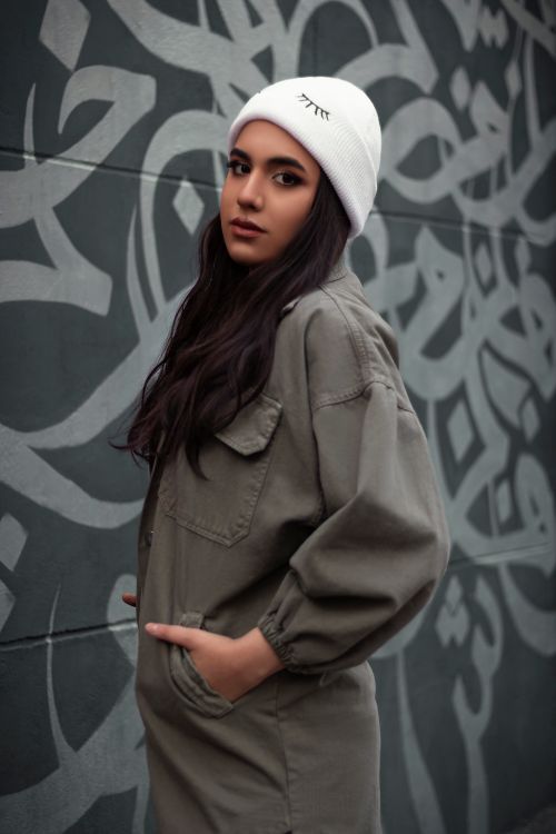 Free Stylish woman wearing a beanie and gray coat with hands in pockets, standing by a graffiti wall. Stock Photo