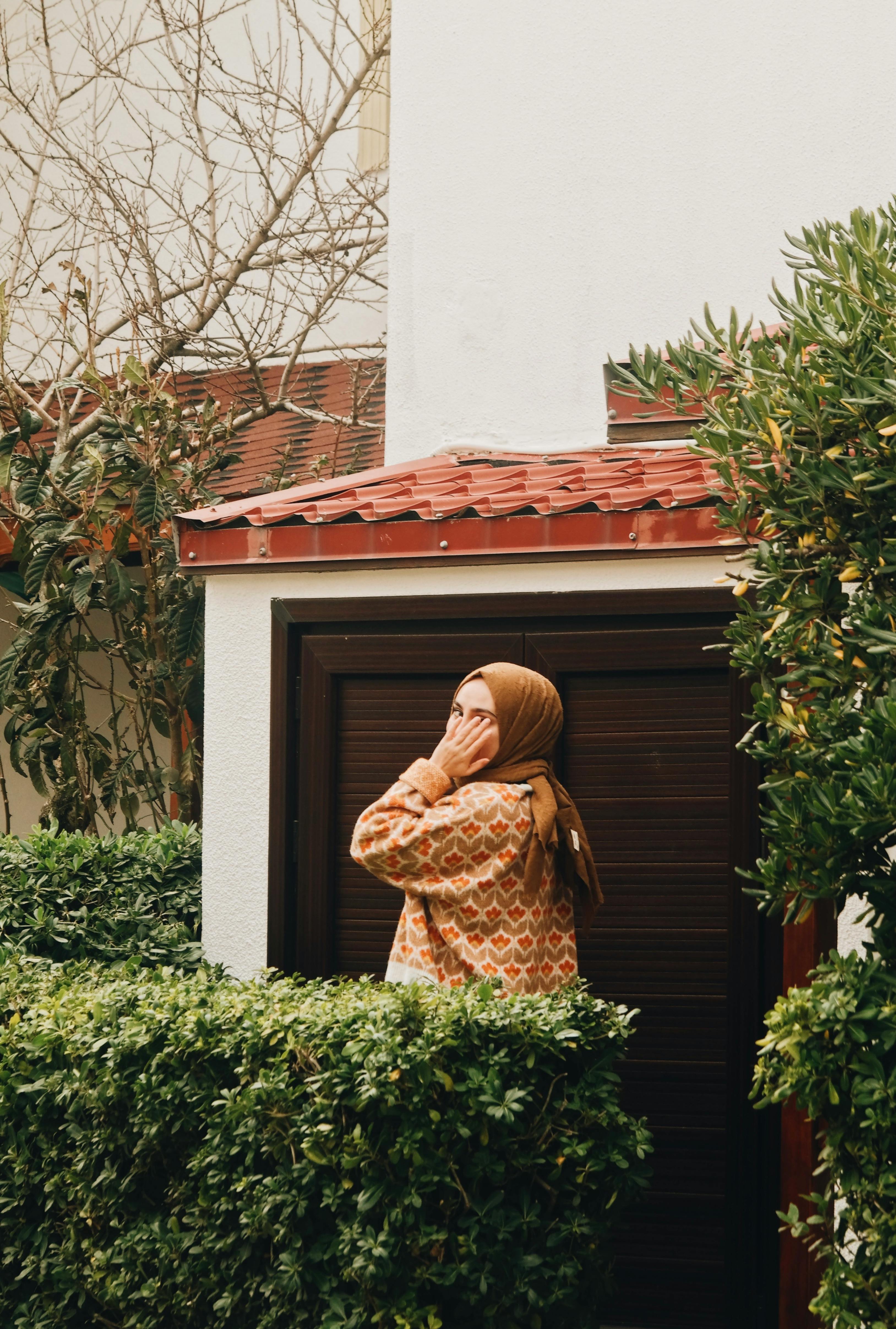 A woman in a printed shirt reflects in a lush garden wearing a headscarf.