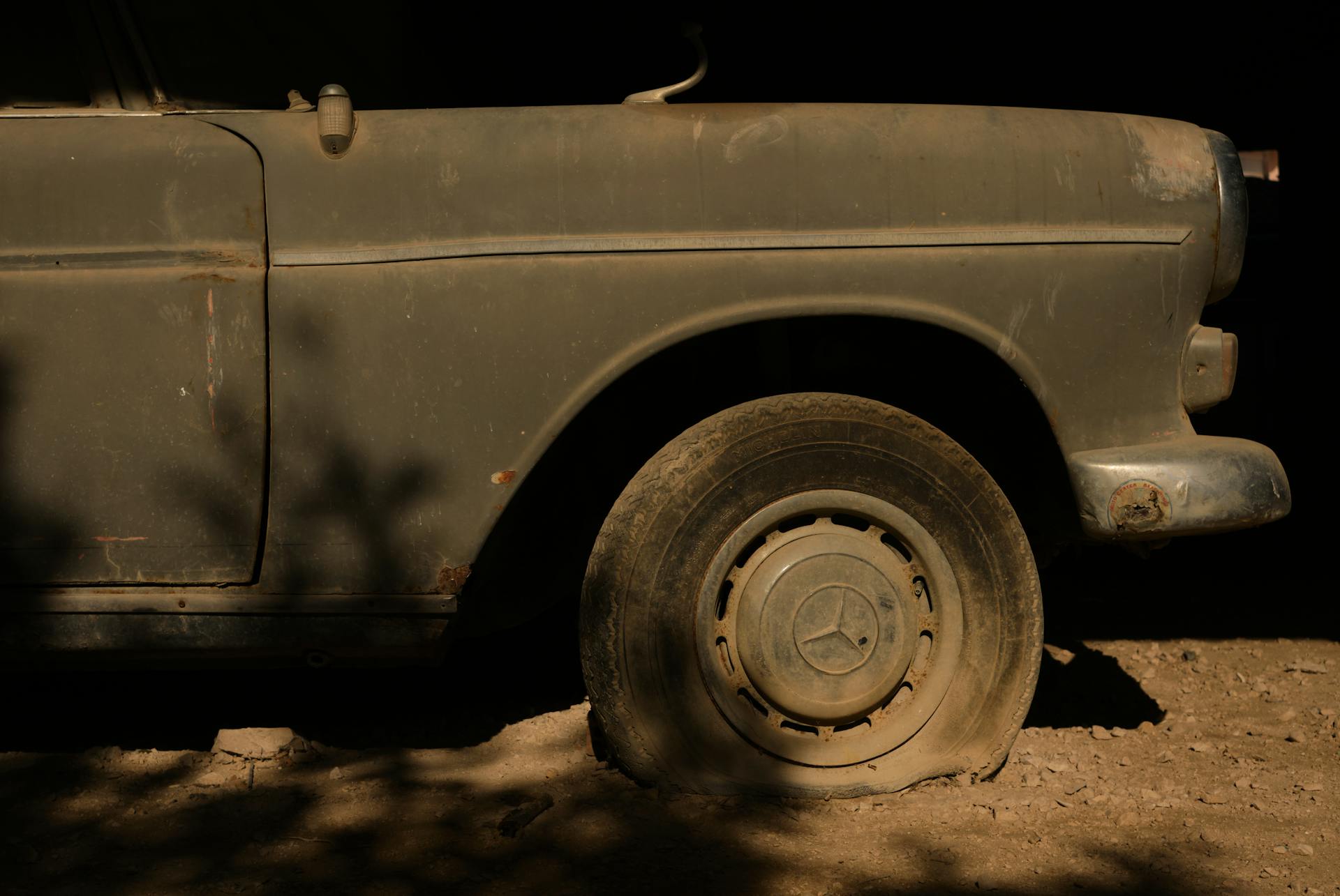 Close-up of the side of a vintage car showing rust and a flat tire under soft light.