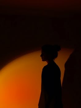A moody silhouette of a woman in profile against a vibrant orange light creating a dramatic shadow effect.