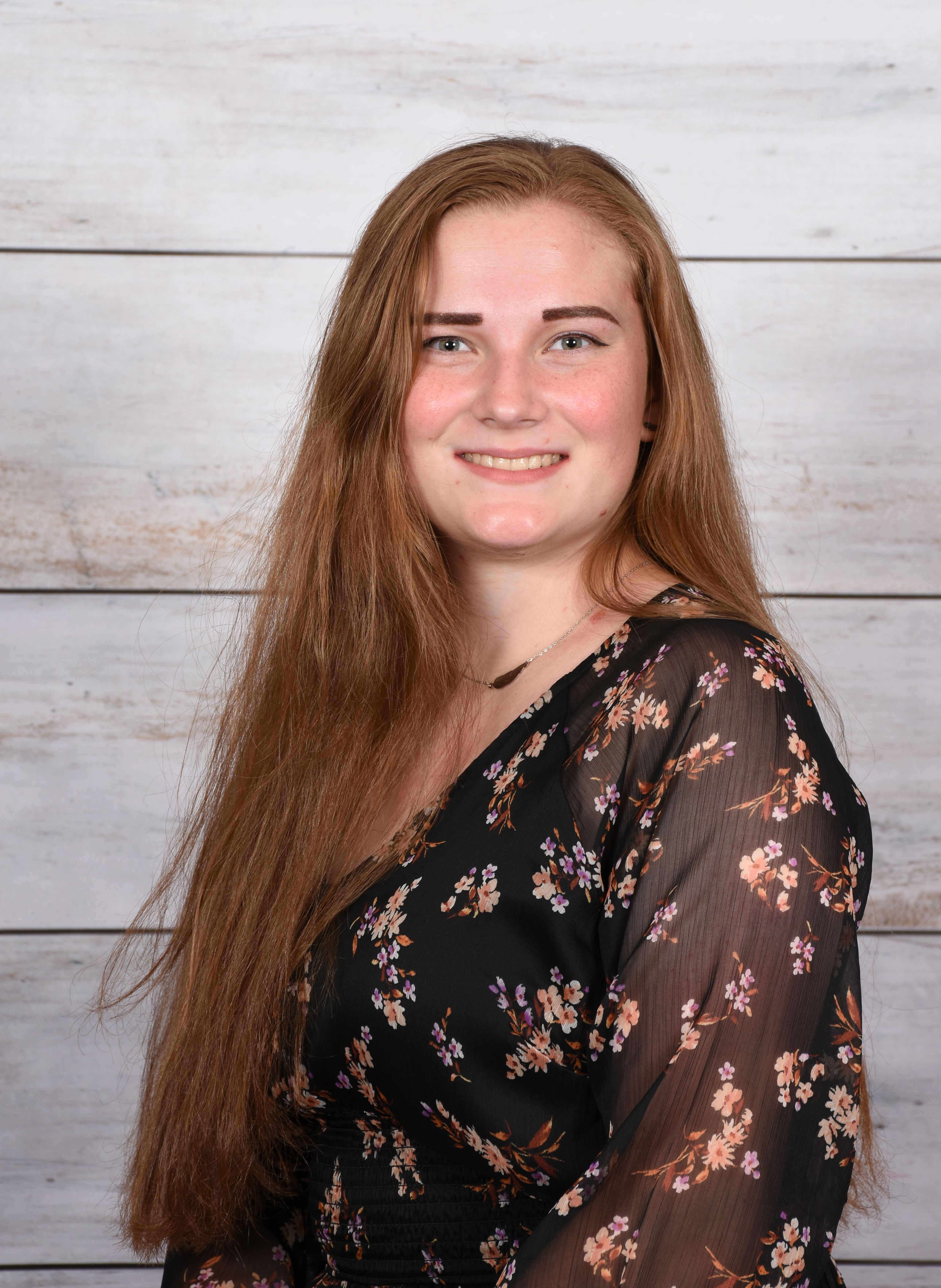 A cheerful woman with long hair in a floral dress poses against a wooden backdrop.
