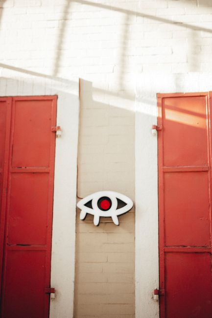 A creative abstract eye sign on a brick wall flanked by red shutters in natural light.
