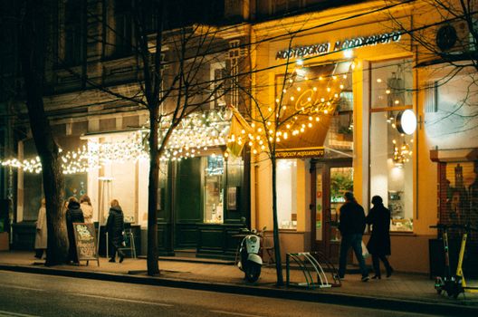 A cozy city street at night with glowing cafe lights and people walking by, creating a warm ambiance.
