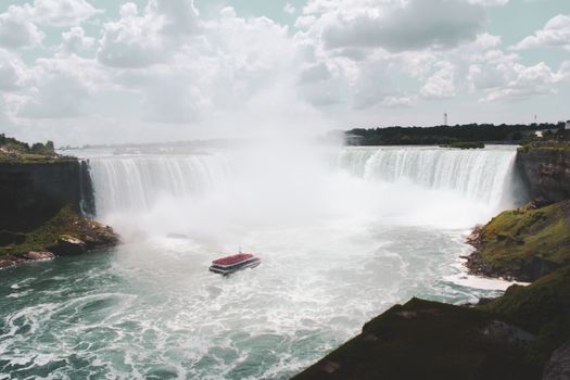 Stunning summer view of Niagara Falls with tour boat and mist under cloudy sky.