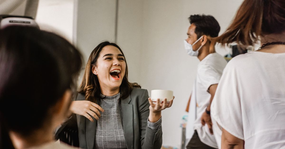 A woman in a gray blazer shares a laugh with colleagues in a lively office setting.