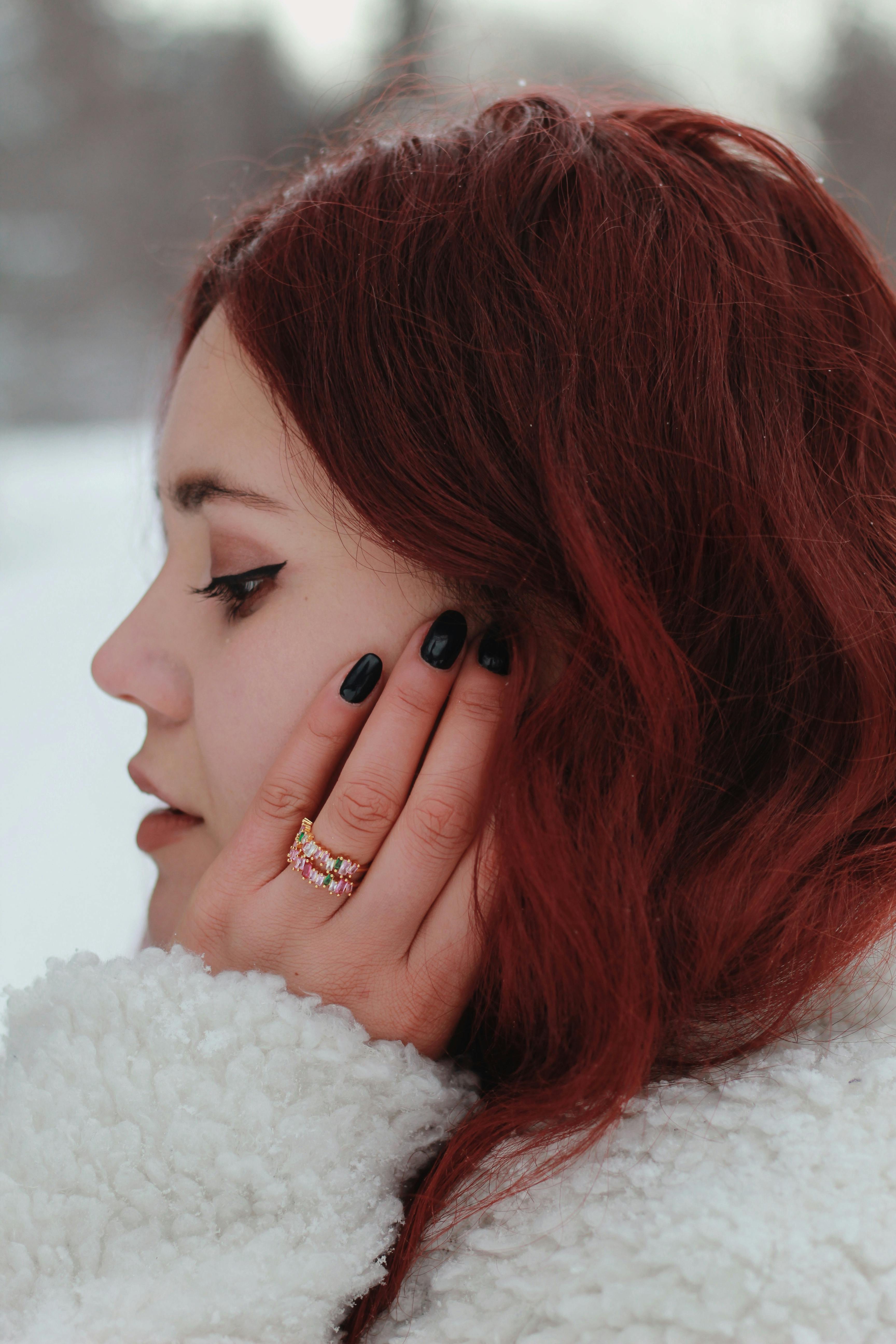 Close-up side profile of a woman with red hair in a winter setting, showcasing rings and black nails.