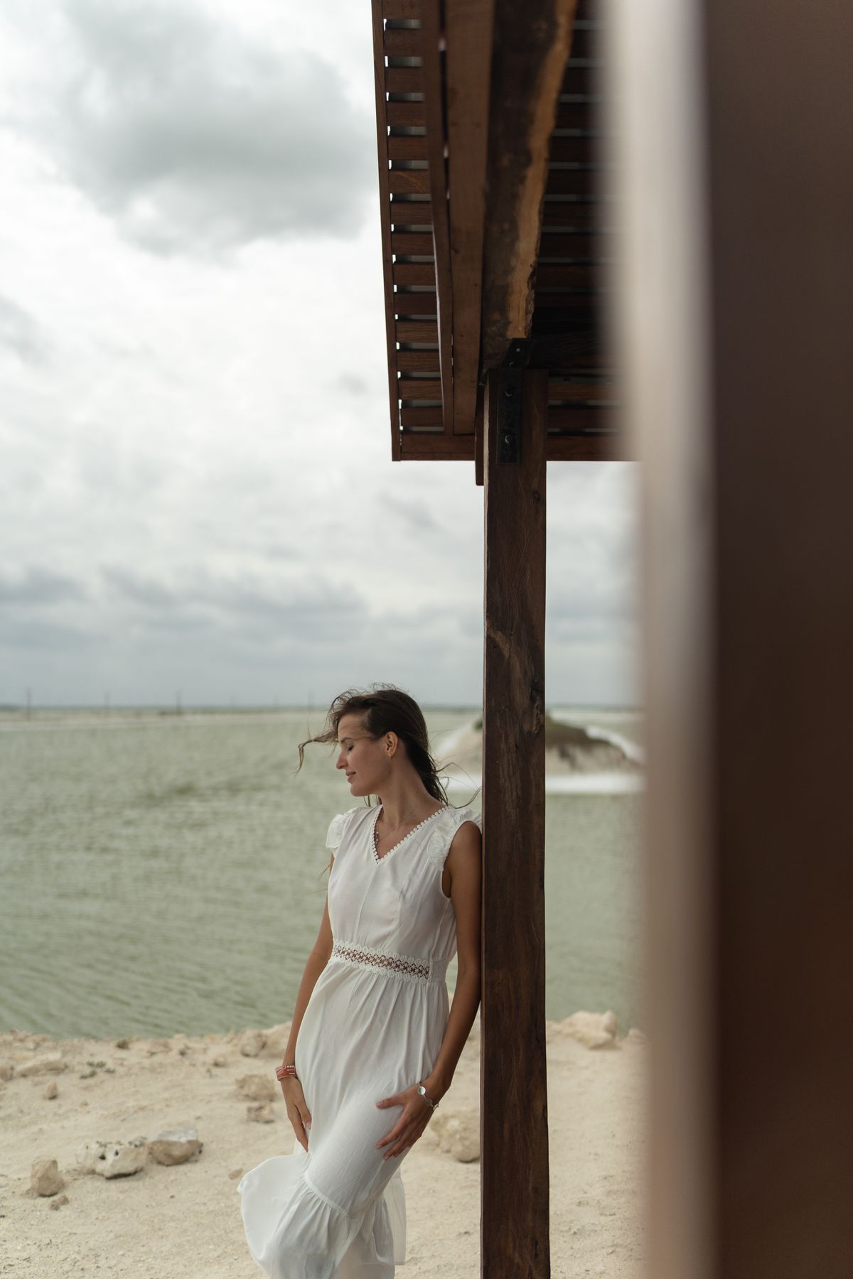 Woman in a white linen dress leaning on a wooden post near a beach
