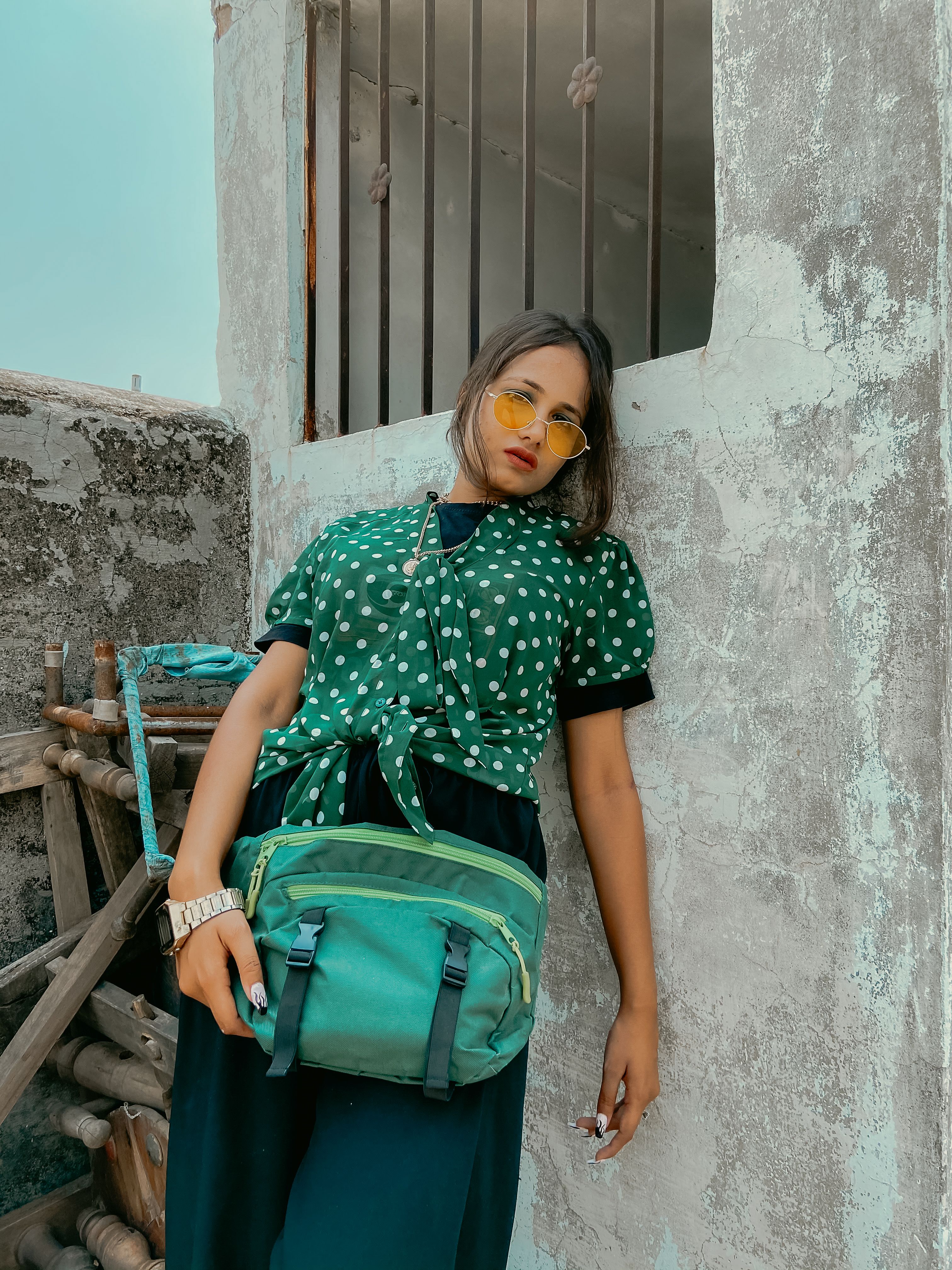 Stylish woman in polka dot blouse leaning against a wall, wearing sunglasses and a turquoise bag.