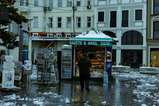 A man approaches a snow-covered newspaper kiosk on a wet winter day in the city.