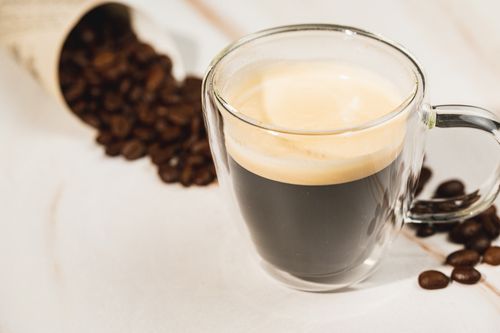 Free A clear glass cup of espresso on a white surface with scattered coffee beans. Stock Photo