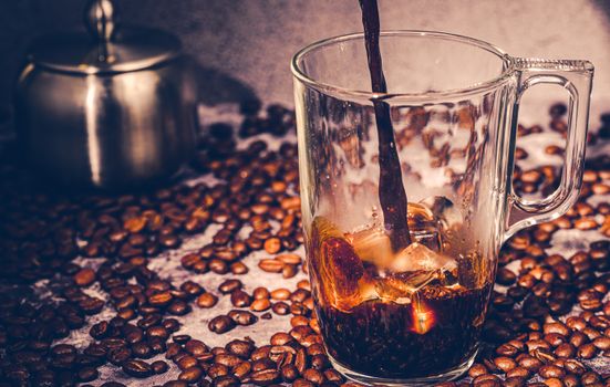 Glass mug of iced coffee being poured over a bed of scattered roasted beans.