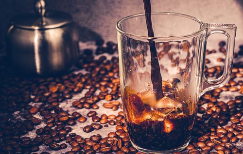 Free Glass mug of iced coffee being poured over a bed of scattered roasted beans. Stock Photo