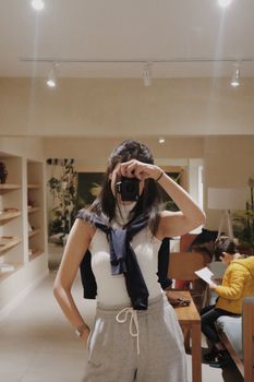 Woman in casual outfit takes mirror selfie indoors, surrounded by modern decor and casual ambience.