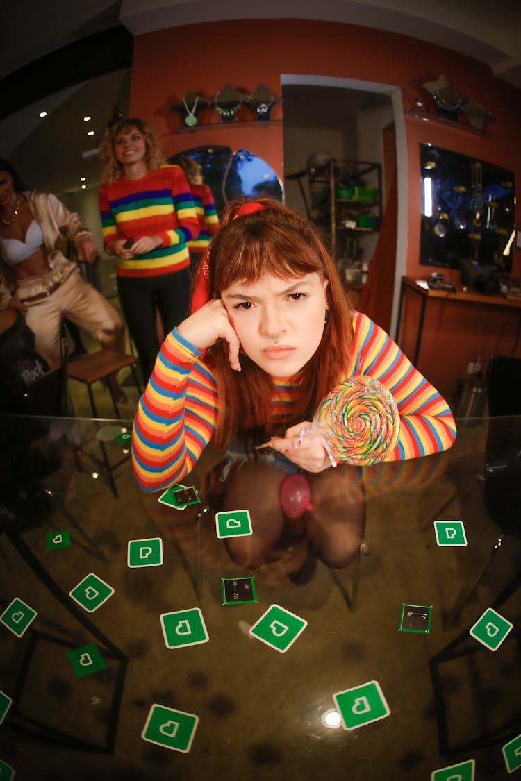 A Young Woman Holding A Rainbow Lollipop Swirl Leaning On Glass Table With Pexels Logo And Pins