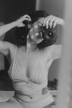 Black and white portrait of a woman taking a selfie in lingerie with a DSLR camera indoors.