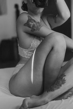 Artistic black and white photo of a tattooed woman in lingerie sitting on a bed.