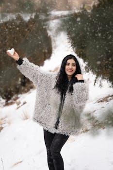 A woman in a fur coat holding a heart-shaped snowball, smiling amidst a snowy landscape.