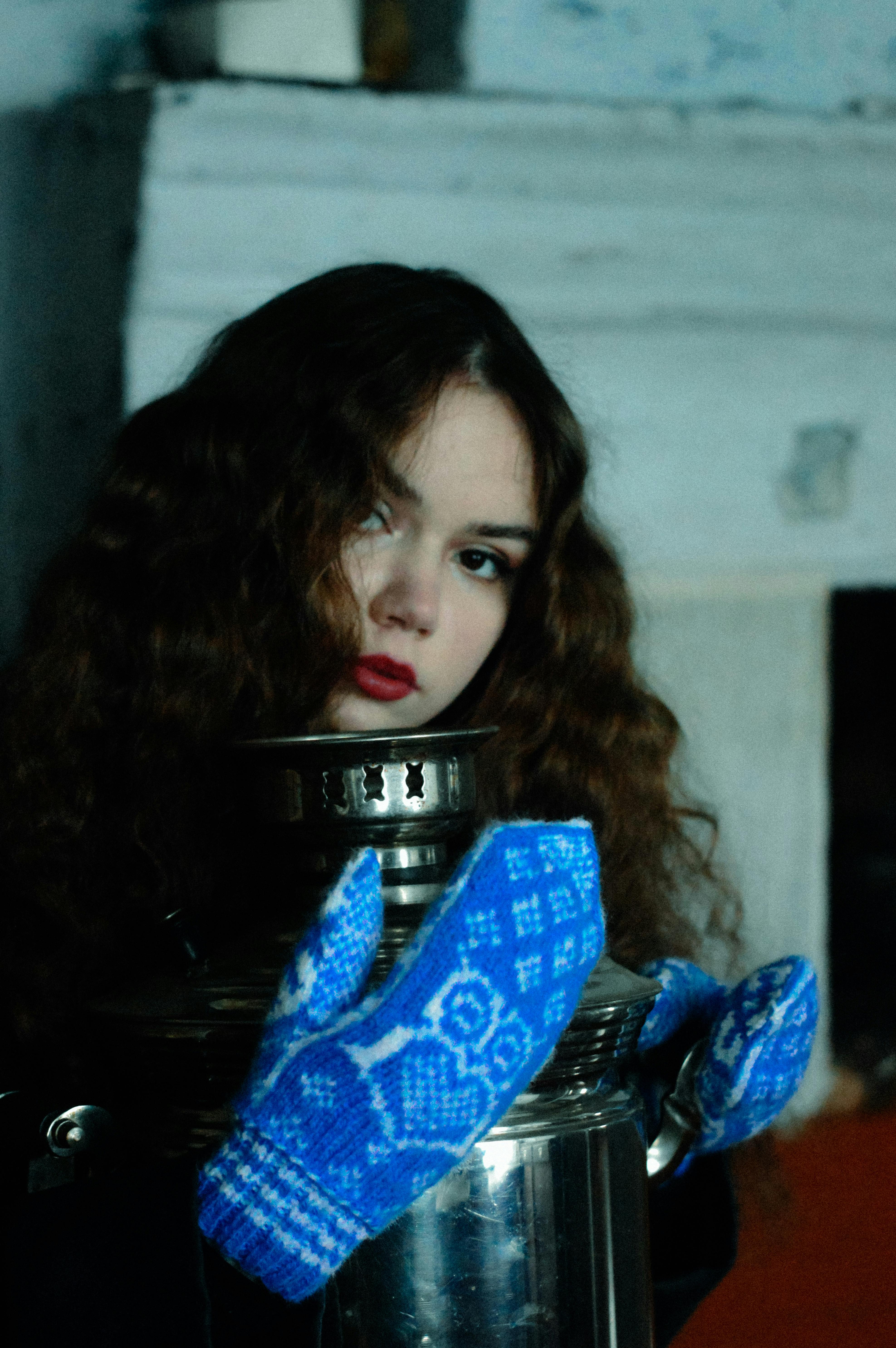 A young woman with curly hair wearing warm clothing, holding a samovar, with blue mittens, in a winter-themed indoor setting.