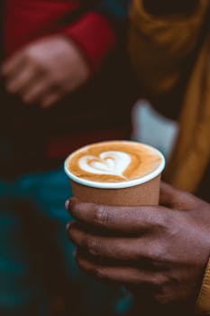 Hand holding a cup of coffee with heart latte art, showcasing barista skill.