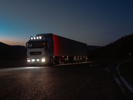 A heavy-duty truck navigates a quiet road at dusk, highlighting its illuminated path through remote mountains.