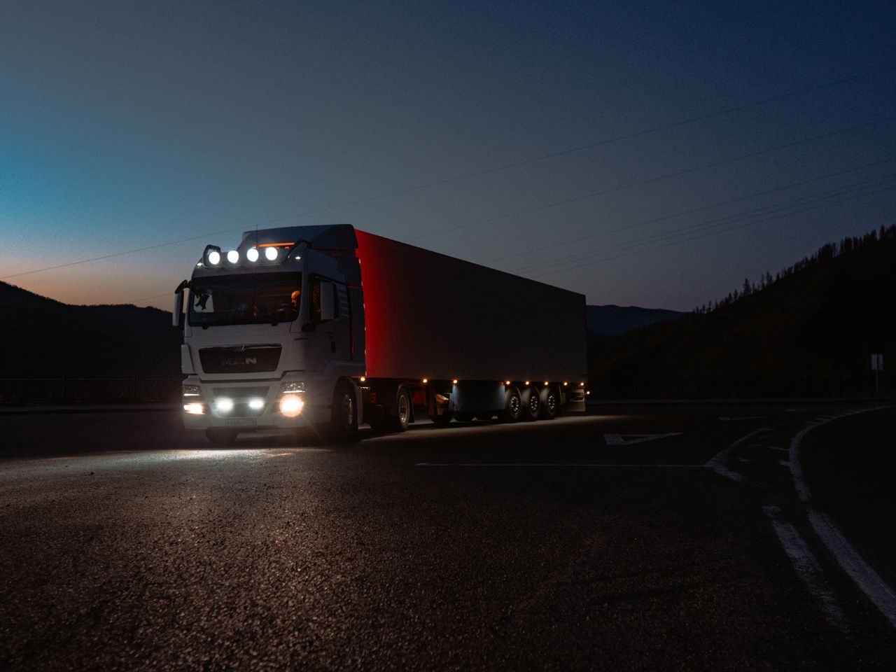An articulated lorry with its headlights on driving along a road at dusk