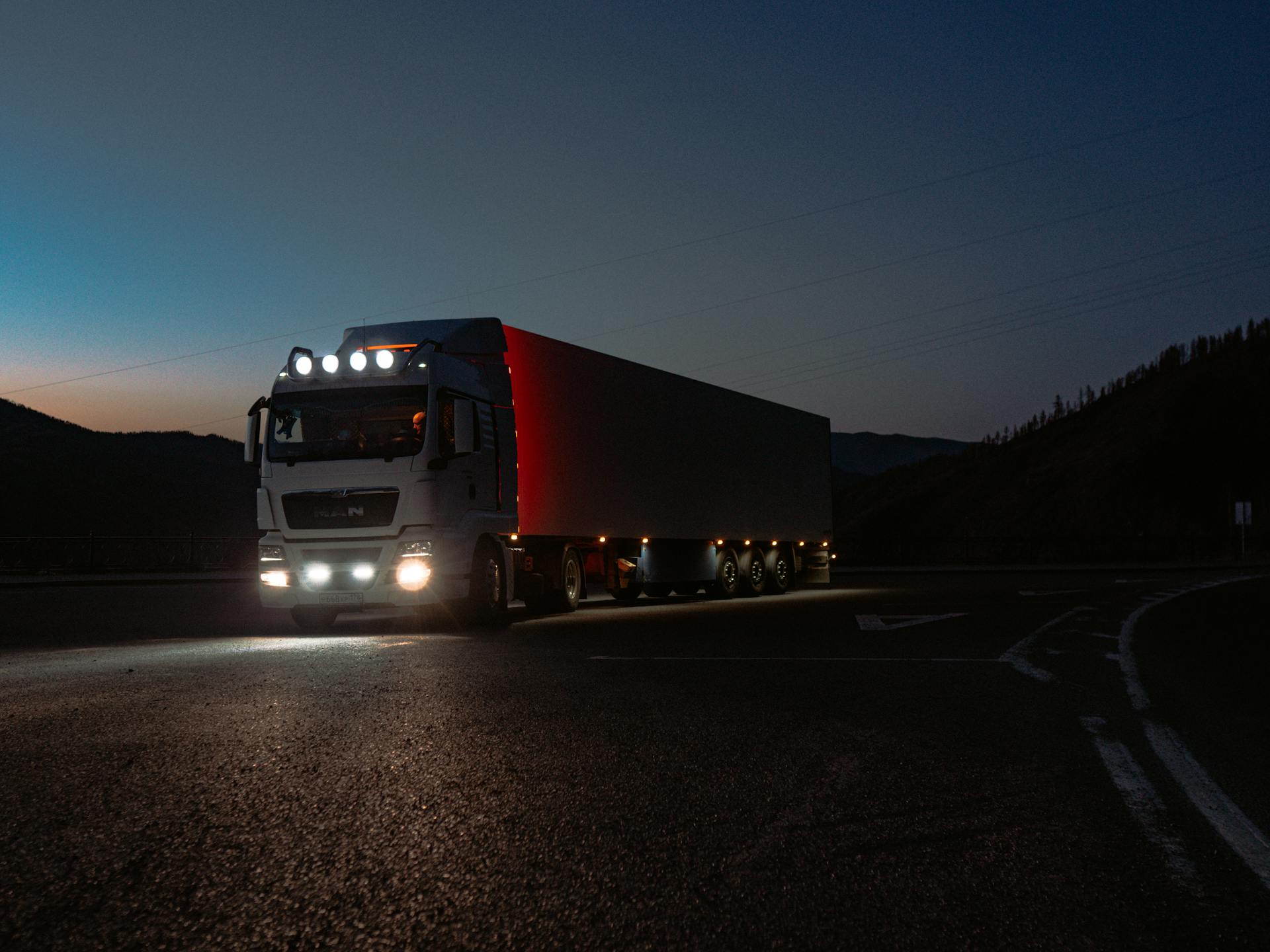 A heavy-duty truck navigates a quiet road at dusk, highlighting its illuminated path through remote mountains.