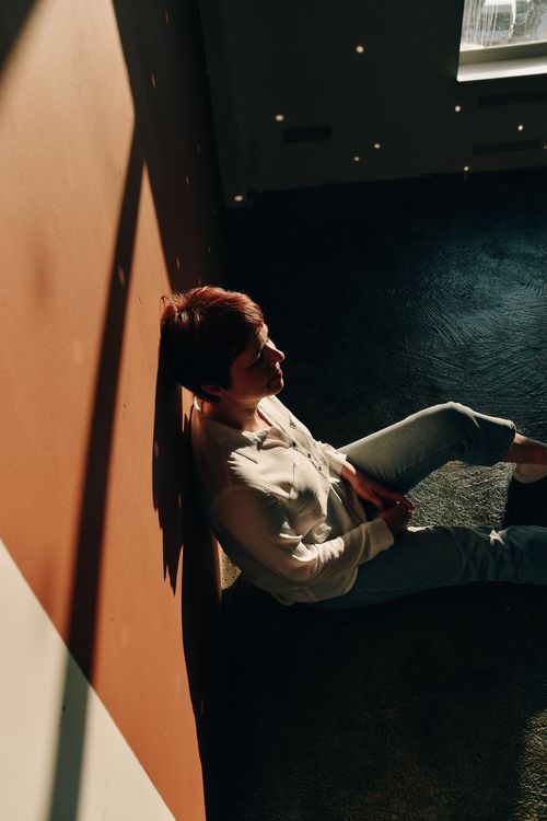 Free A woman with short red hair sits thoughtfully against a warmly lit wall indoors. Stock Photo