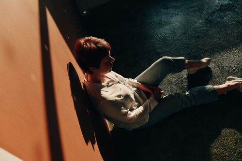 Free A woman wearing white long sleeves sits against a wall, bathed in sunlight, creating interesting shadows. Stock Photo