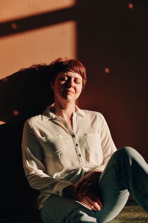 Free A woman with short hair sits against a wall, enjoying warm sunlight on her face. Stock Photo