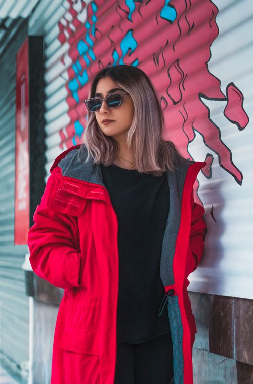 Free Stylish woman wearing a red jacket and sunglasses poses against a colorful urban background. Stock Photo