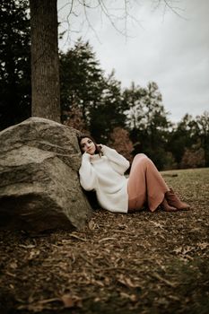 Woman in a sweater sitting by a rock in a serene fall landscape.