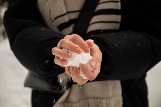 A person wearing a coat and scarf holds a snowball outdoors during winter.