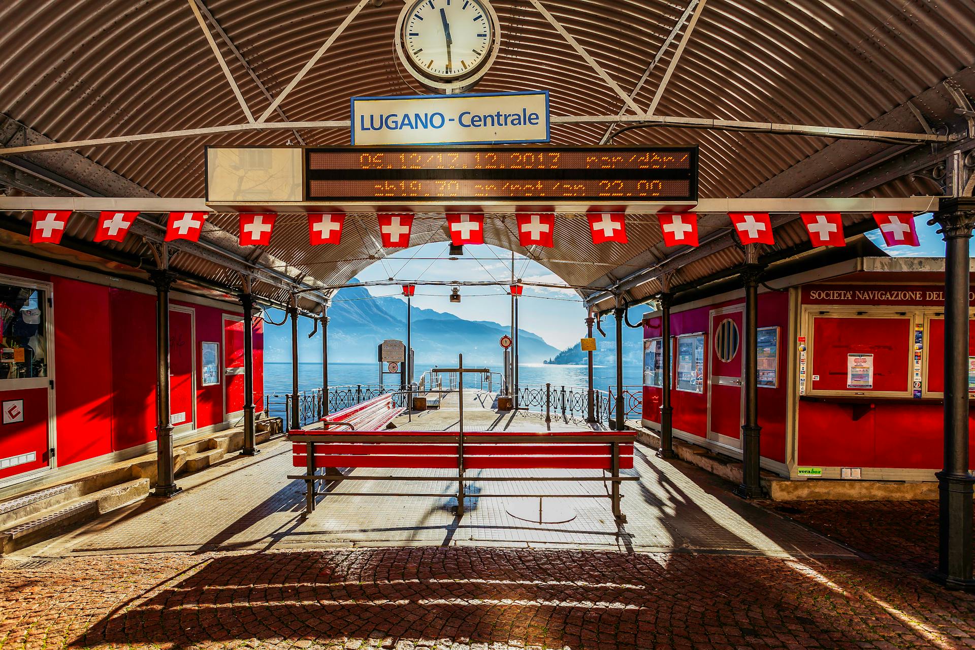 A bright day at Lugano Centrale station with Swiss flags and lake view.