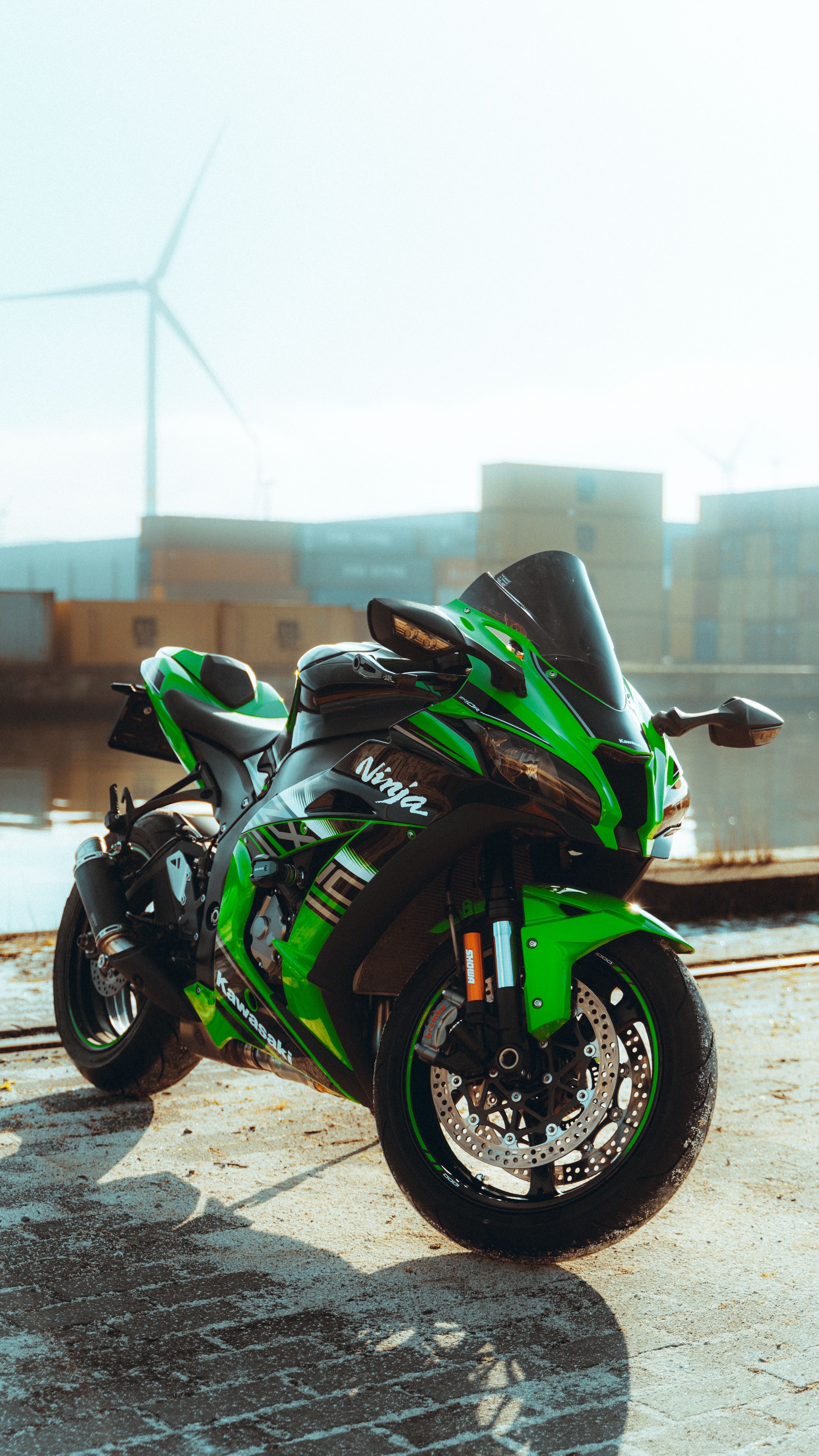 A vibrant green sports motorcycle parked by a waterfront with wind turbines in the background.