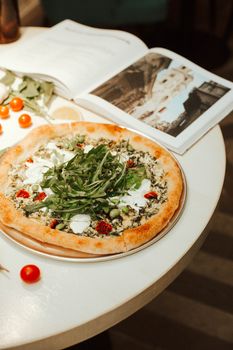 Gourmet pizza topped with arugula and ricotta beside an open book on a cafe table.