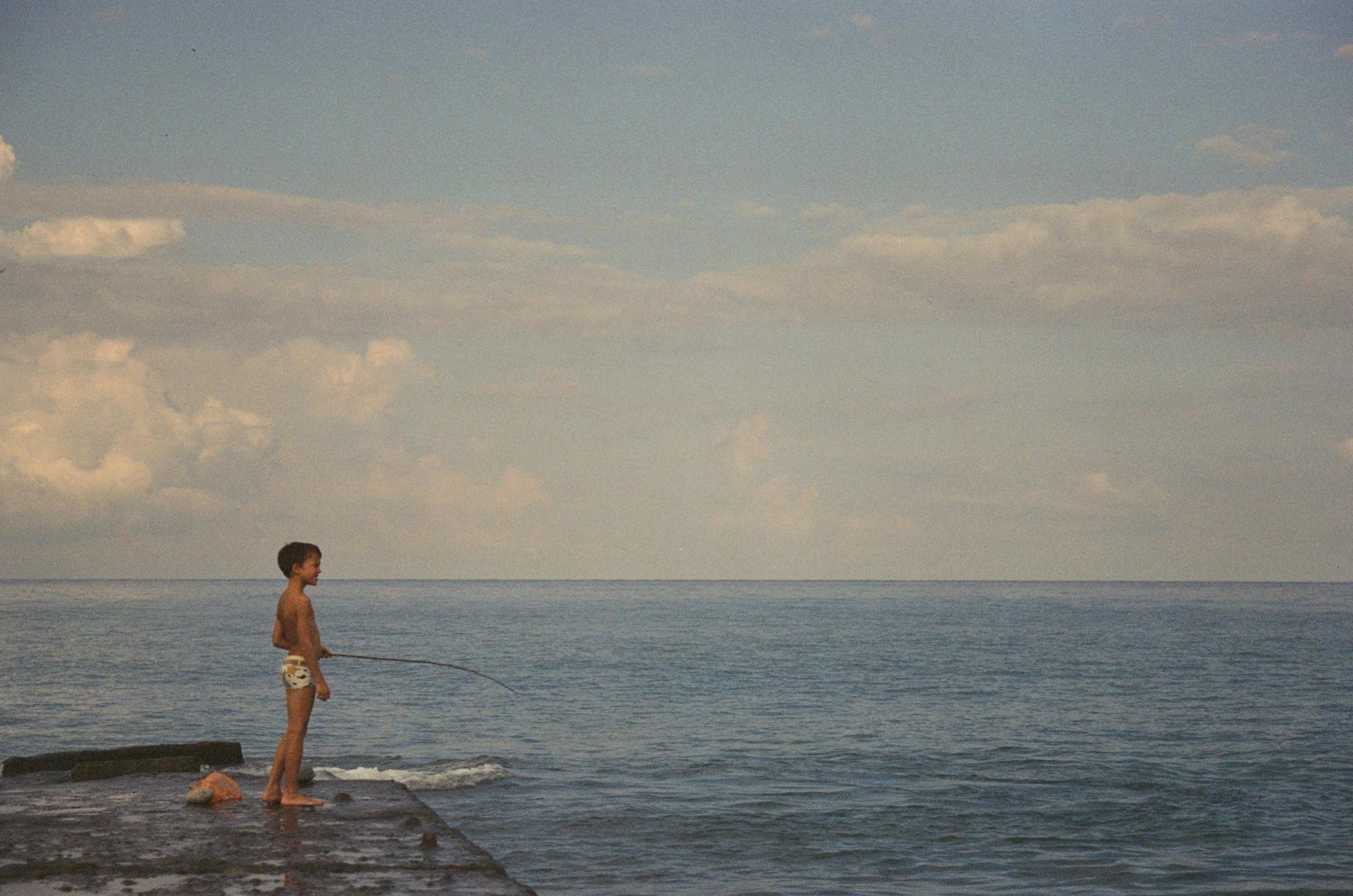 A young boy fishing on a pier by the sea in Adler, Russia, under a cloudy sky.
