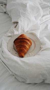 Freshly baked croissant served on a decorative white plate, placed on a cozy bed.