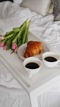 A cozy morning scene with coffee, croissant, and tulips on a bed tray, enhancing relaxation.