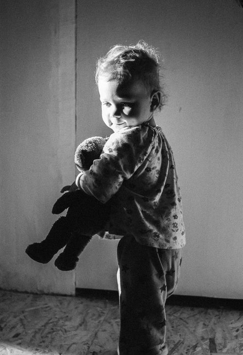 Free A child stands indoors holding a teddy bear, highlighted by dramatic lighting. Stock Photo