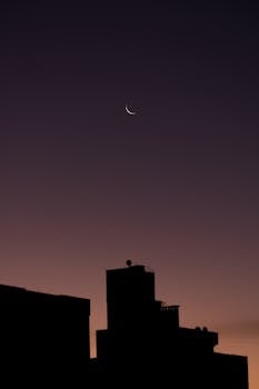 A city skyline silhouette with a crescent moon during twilight, creating a dramatic urban scene.
