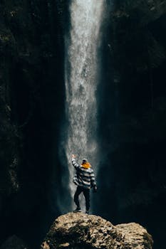 A person in a knit hat stands on a rock, admiring a powerful waterfall in a natural setting.