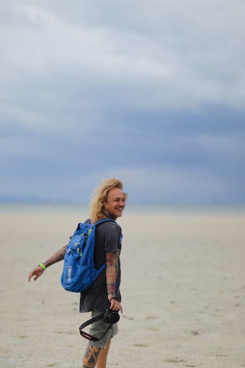 Free A smiling man with tattoos and a camera backpack exploring a vast desert. Stock Photo