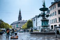 A street musician plays guitar by a fountain in Copenhagen city center, surrounded by urban architecture and pedestrians.