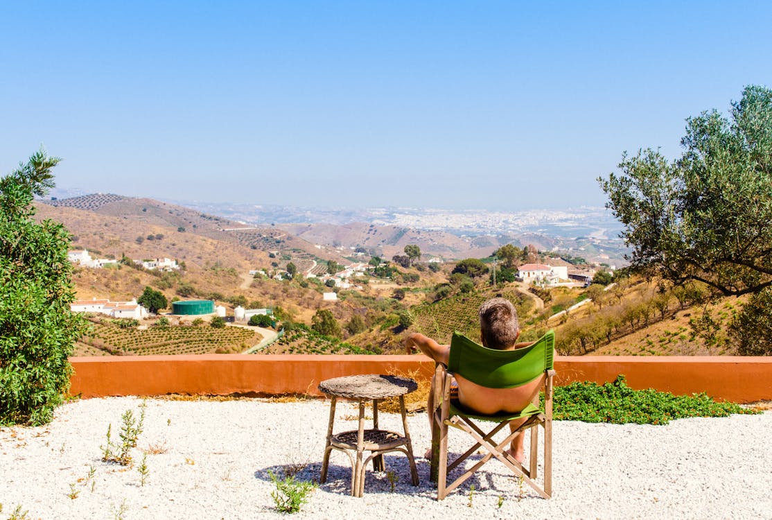 Free Man Sitting on Green Chair Near Trees and Mountain Under Blue Sky at Daytime Stock Photo