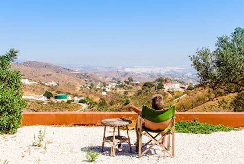Man Sitting on Green Chair Near Trees and Mountain Under Blue Sky at Daytime