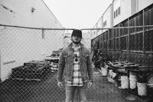 Grayscale Photo of Man in Denim Jacket Standing Near Fence
