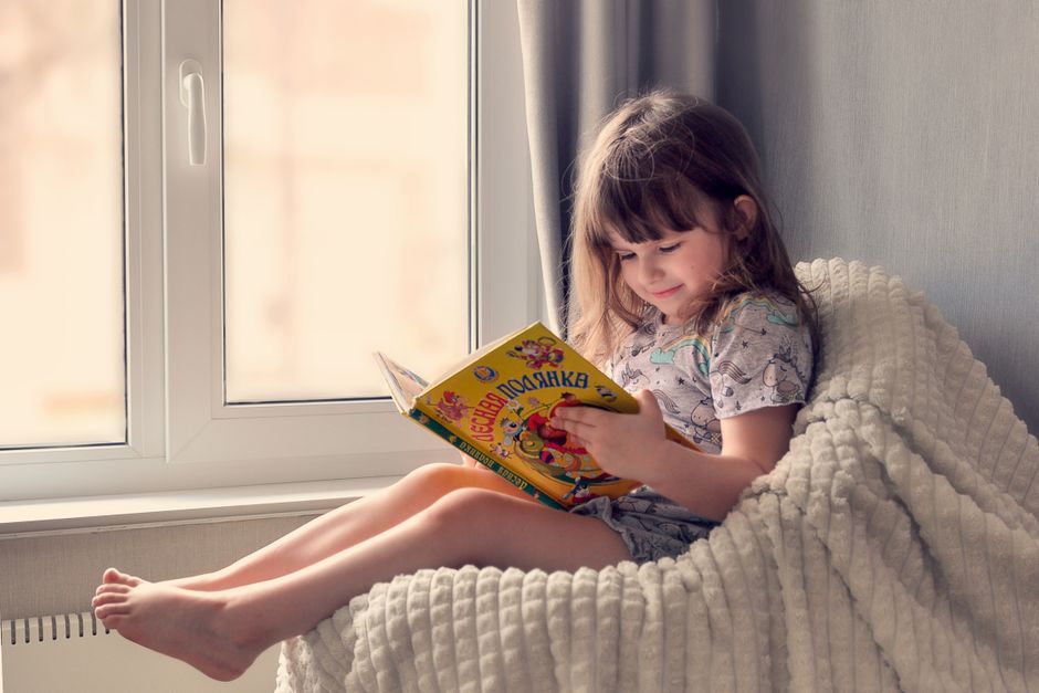 Photo by Ольга Жарикова on Pexels A young girl reads a colorful book by the window, embracing cozy and peaceful leisure time indoors. - Photo by Ольга Жарикова on Pexels