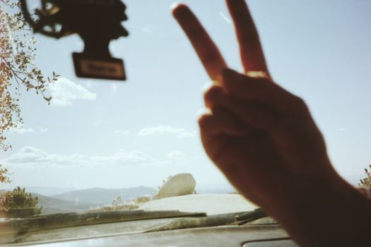 A hand makes a peace sign inside a car overlooking a scenic mountain landscape.