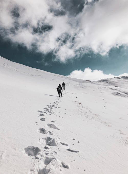 Low-Angle Shot of Two People Walking on Top of Snow-Covered Mountain under the Cloudy Sky