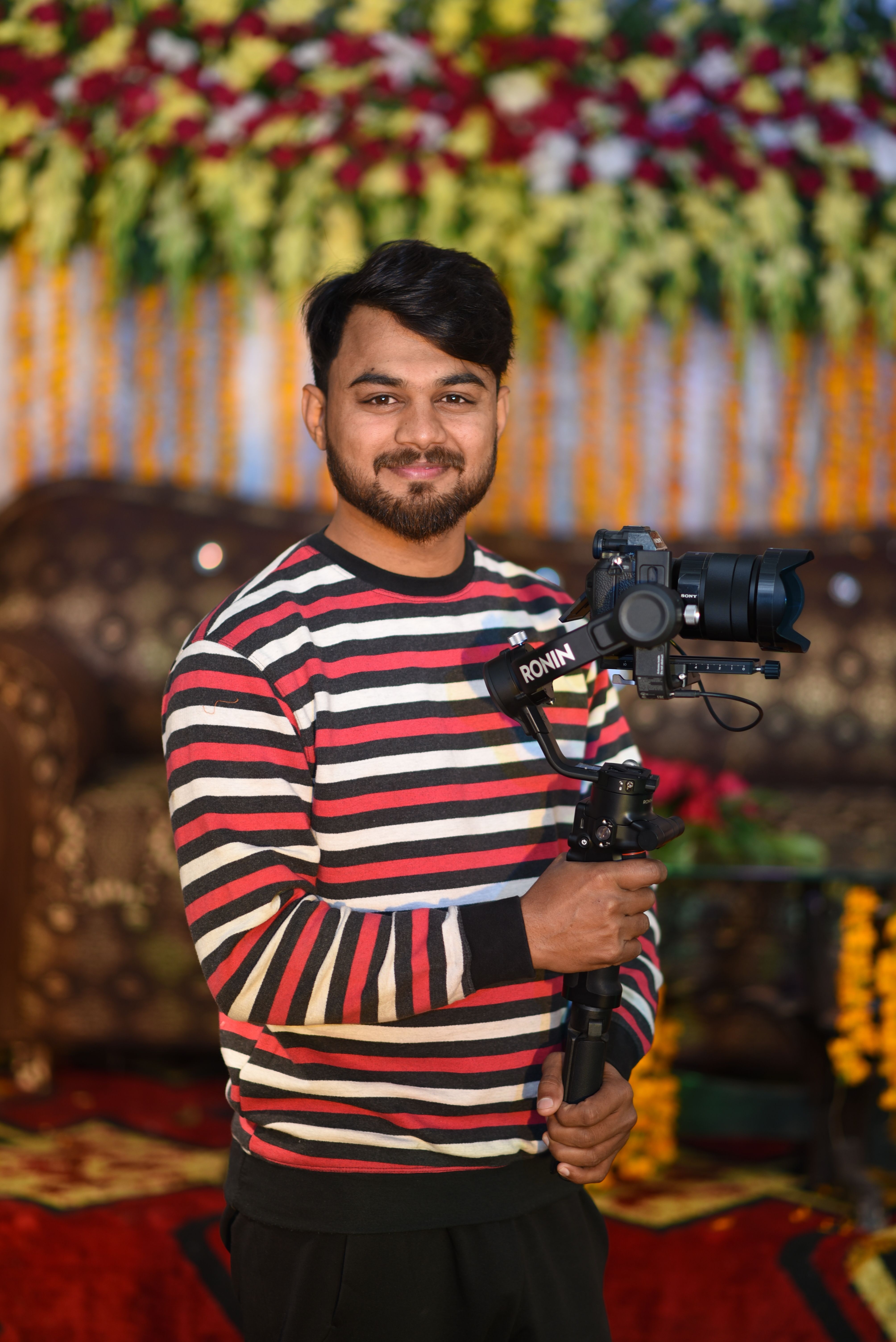 A smiling photographer posing indoors with a professional camera, wearing a striped shirt and holding a stabilizer.